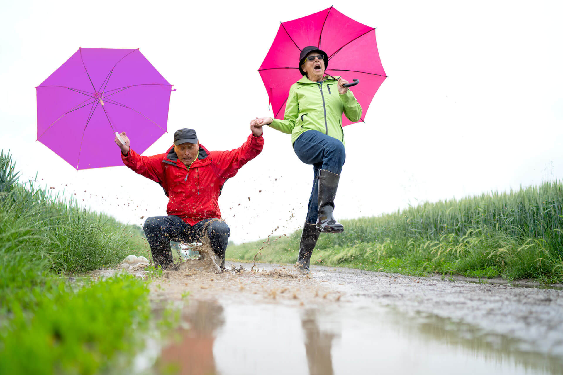 funny active 70 years old senior couple hand in hand on path through rural landscape on bad weathers day, man jumping in dirty rain puddle